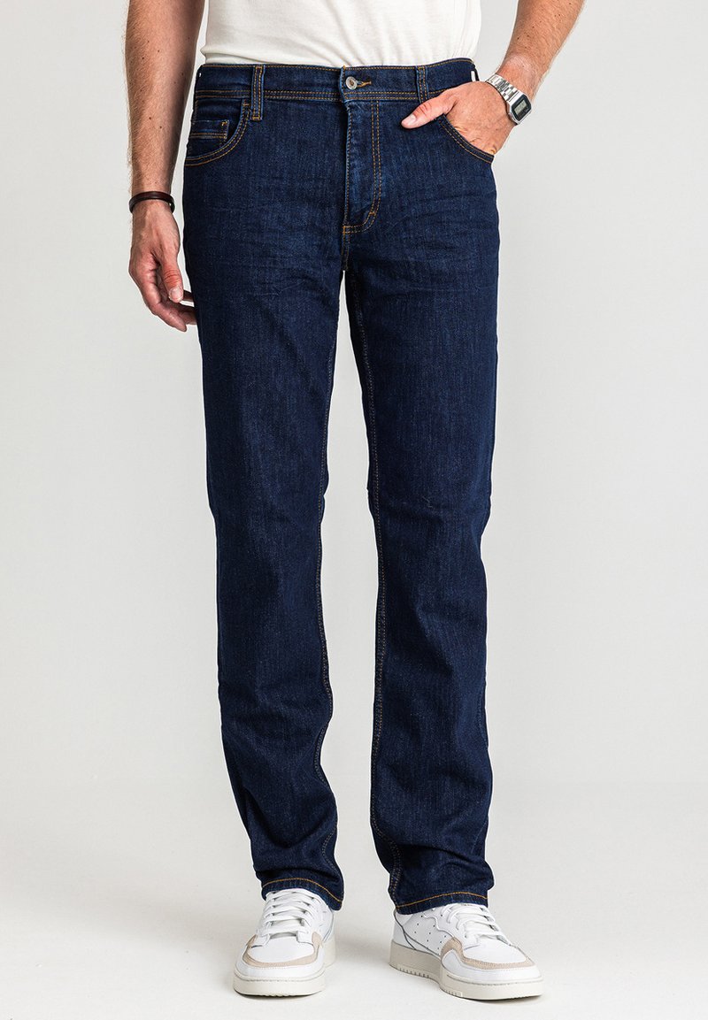 Ciemnoniebieskie jeansy z denimu o prostym kroju nogawek, z żółtym przeszyciem i przednimi kieszeniami. Noszone z białymi tenisówkami i prostą białą koszulą.