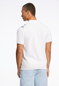 Weißes kurzärmliges Poloshirt mit strukturiertem Finish, das blaue und weiße gestreifte Akzente an den Schultern und einen geraden Saum aufweist.