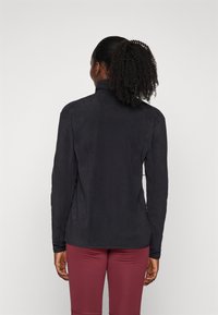 Maglietta nera a maniche lunghe con colletto alto e texture liscia. Presenta una vestibilità elegante e un design minimale. Indossata con leggings bordeaux.