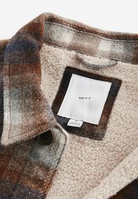 Chemise en flanelle à carreaux de couleurs marron, gris et crème avec une doublure en polaire. Elle présente une étiquette et une fermeture à bouton marron.