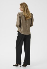 Pikkade blondide juustega naine kannab säravat oliivirohelist pluusi, musti laiadest jalgadest pükse ja musti madala kandadega kingi, olles seljaga pööratud.