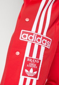 Červená sportovní tkanina Adidas s bílými třemi pruhy, vyšitým logem Adidas a nášivkou s nápisem "Značka se třemi pruhy."