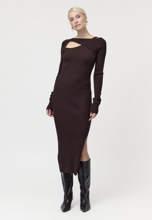 Femme aux cheveux blonds courts portant une robe marron près du corps en côtes avec une découpe et des bottes noires montantes sur fond blanc.