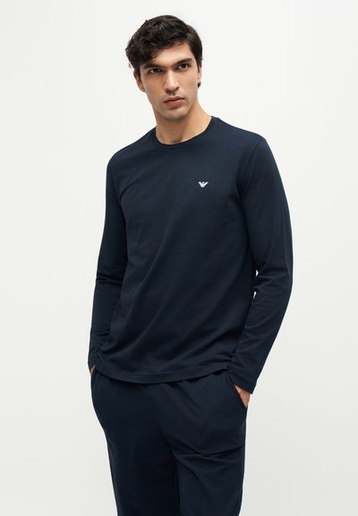 Jeune homme aux cheveux foncés portant une chemise bleu marine à manches longues et un pantalon assorti sur un fond clair uni.