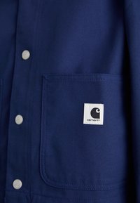 Veste bleu marine en tissu durable, dotée de boutons-pression, de deux poches et d'une étiquette blanche avec un logo noir sur la poitrine gauche.
