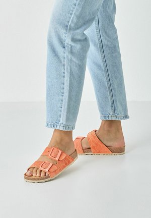 Sandali arancioni con texture, dotati di due fibbie regolabili e suola in sughero, abbinati a jeans di denim azzurro chiaro con risvolto.