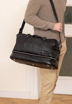 Borsa da viaggio in pelle nera con forma arrotondata, chiusura con zip, due manici superiori e tracolla rimovibile. Presenta piedi in metallo nella parte inferiore.