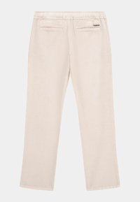 Beige broek met een gladde textuur, elastische tailleband en twee achterzakken. Heeft een klein zwart "GUESS" logo op de tailleband.