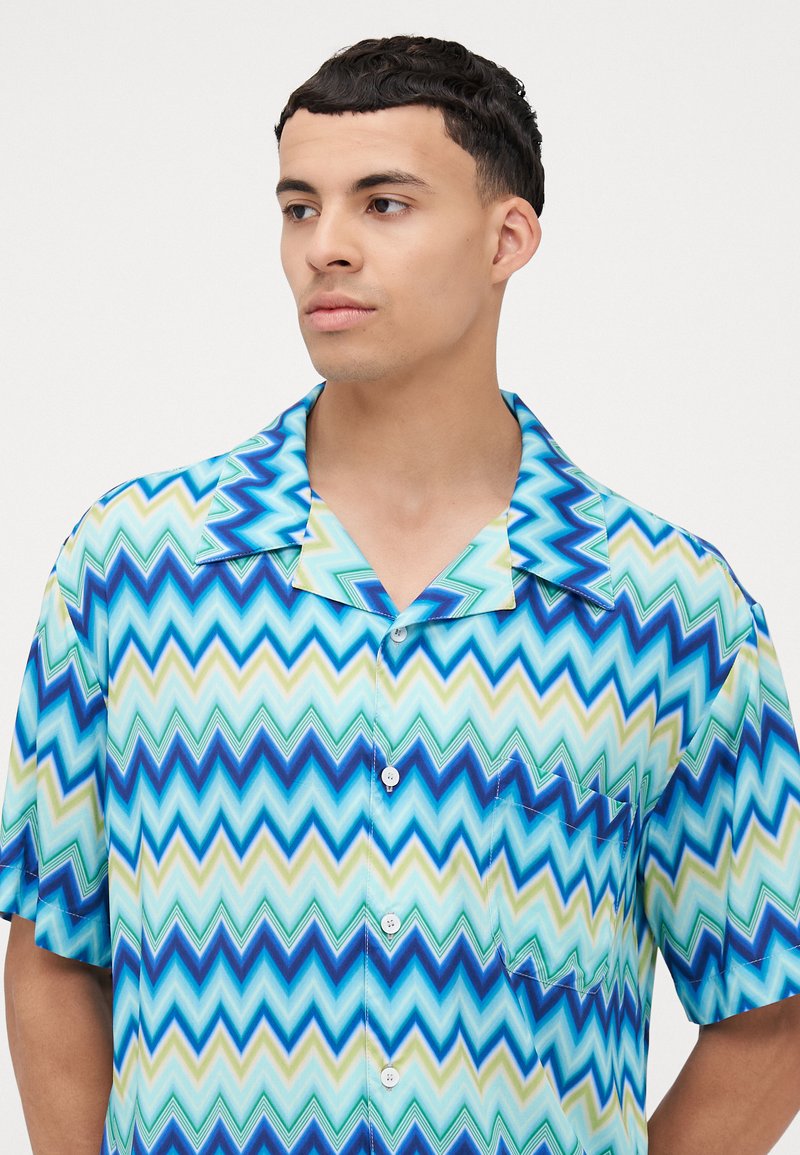 Camicetta a maniche corte in tessuto leggero con un motivo a zigzag nei toni di blu, acquamarina e verde. Include una tasca e chiusura con bottoni.