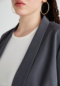 Šedý blazer s hladkou texturou, s klopovým límcem, vrstvený přes bílou žebrovanou košili. Náušnice ve tvaru kruhu přidávají metalický akcent.