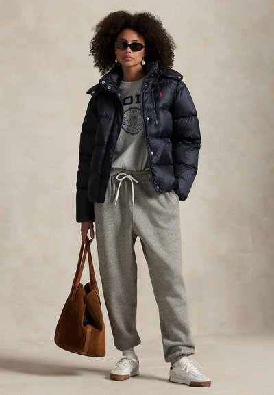 Vrouw draagt een zwarte pufferjas, grijze sweatpants en sweatshirt, witte sneakers, zonnebril, houdt grote bruine suède tas vast.