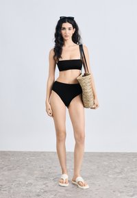 Σετ μαύρου μπικίνι με τοπ μπαντέο και σλιπ με ψηλή μέση, σε συνδυασμό με πλεκτή ψάθινη τσάντα και λευκές σαγιονάρες. Λεία υφή.