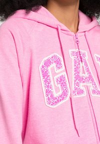Sudadera de color rosa con cierre, que presenta un gran logo "GAP" en un tono rosa más claro. El material es suave y tiene una textura lisa.
