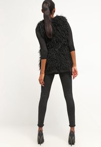 Svart fuskpälsväst med en strukturerad yta, kombinerad med svarta skinny jeans. Modellen bär svarta högklackade skor.