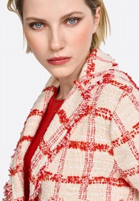 Veste en tweed blanche avec un motif à carreaux rouges, bords frangés et revers. Portée sur un haut en tricot rouge. Cheveux courts et yeux bleus visibles.