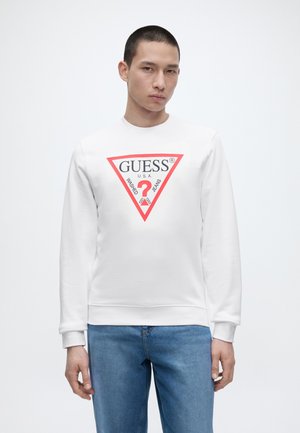 Mann trägt weißen Sweatshirt mit rotem und schwarzem GUESS-Logo und blauen Jeans, steht vor weißem Hintergrund.