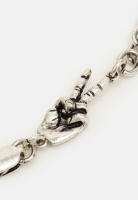 Pulseira de metal com tom prateado, apresentando um pendente em forma de mão fazendo o gesto de paz, com detalhes nos nós dos dedos e articulações.