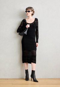 Abito nero lavorato a maglia con maniche lunghe e un motivo testurizzato, abbinato a stivaletti neri a punta e una borsa nera.