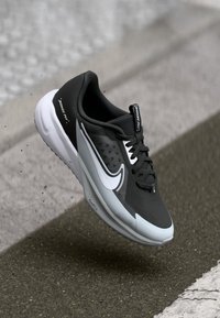 Fekete futócipő textúrált felsőrésszel, fehér swoosh logóval, szürke középtalppal és fekete fűzőkkel. Párnázott boka része és gumi talpa van.