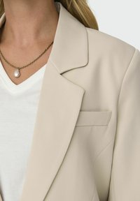 Blazer beige realizzato in tessuto texturizzato, con rever a scollatura e una singola tasca sul petto. Indossato sopra una camicia bianca a scollo a V.