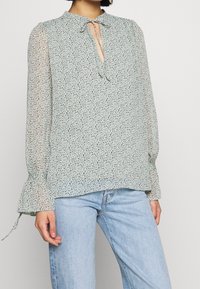 Ljusgrön blus med blommönster, nyckelhålsknut, långa genomskinliga ärmar med rynkade manschetter och en lös passform.