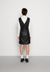 Schwarzes Kunstlederkleid mit V-Ausschnitt, ärmellos und knielang. Kombiniert mit weißen Socken und schwarzen Schnürschuhen.