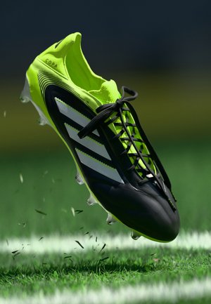 Black and neon yellow soccer cleat with three white stripes, textured upper, black laces, and clear studs, appearing above green grass.