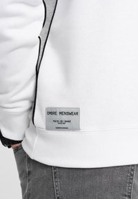 Weißer Kapuzenpullover mit grauem Akzent, geripptem Saum und einem Etikett mit der Aufschrift "OMBRA MENSWEAR" an der Seite, was auf europäisches Design hinweist.