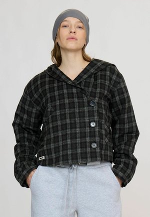 Femme portant un bonnet gris, une veste à carreaux noir et gris boutonnée avec un grand col, et un pantalon de survêtement gris clair avec les mains dans les poches.