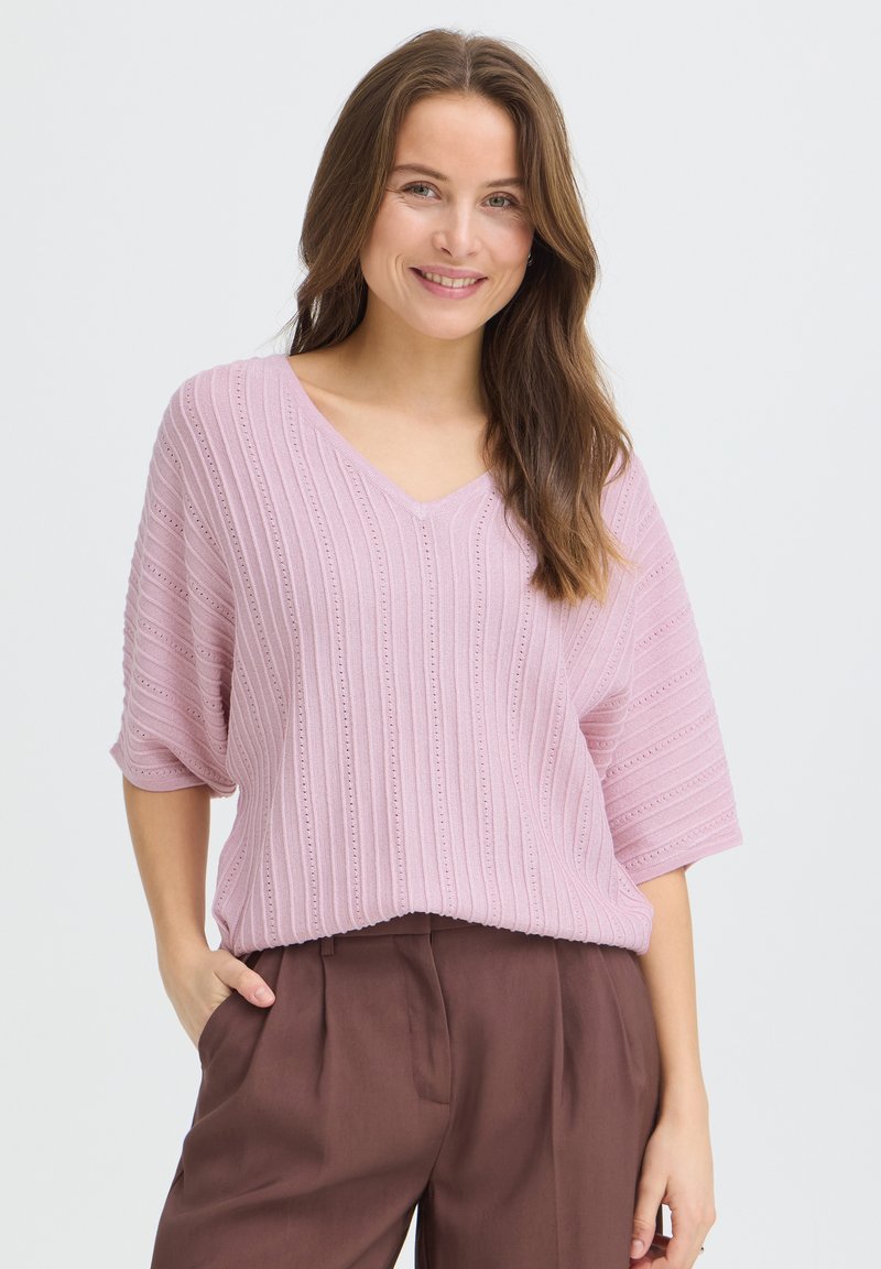 Femme aux longs cheveux bruns souriante, portant un haut en maille rose clair à col en V et un pantalon marron taille haute, la main dans la poche.