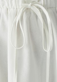 Fascia in tessuto bianco con texture arricciata, dotata di due lunghi nastri. Il materiale è leggero e leggermente traslucido, con una finitura liscia.