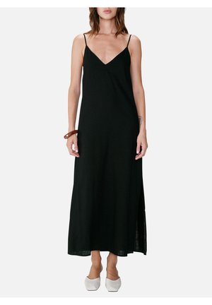 Kvinde iført en lang sort ærmeløs kjole med tynde stropper, hvide sko med firkantet tå og en brun armbånd på venstre håndled.