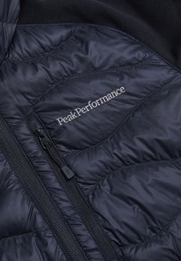 Marine geïsoleerde jas met een geplooide textuur, voorzien van een ritssluiting aan de voorkant, PeakPerformance-logo en gladde stoffen accenten.