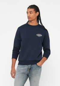 Mørkeblå genser med rund hals og ribbestrikkede mansjetter, med en liten hvit logo på brystet. Kombinert med lyseblå jeans.