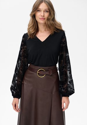 Zwarte top met V-hals en doorzichtige, geborduurde lange mouwen, gecombineerd met een bruine leren rok met een brede gouden cirkelvormige riemgesp.