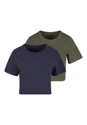 Femme aux cheveux bouclés portant un t-shirt court bleu marine et un pantalon beige à pois, debout avec une main sur la hanche.