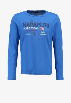Homme portant une chemise bleue à manches longues, un jean bleu foncé, des baskets grises et un sac à dos blanc avec une sangle, debout avec une main dans la poche.