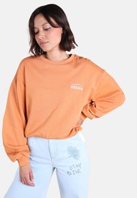 Sweatshirt orange à manches longues avec un col rond et un logo "OXBOW" brodé en blanc. Assorti à un jean bleu clair avec des broderies florales et textuelles.