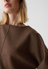 Bruine trui met een losse pasvorm, voorzien van een brede halslijn en glad, gestructureerd stof. Eenvoudige, strakke lijnen zonder zichtbare bevestigingsmaterialen.