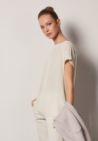 Ljus beige, satinlik blouse med korta ärmar och rund halsringning. Har subtila veck och en slät yta.