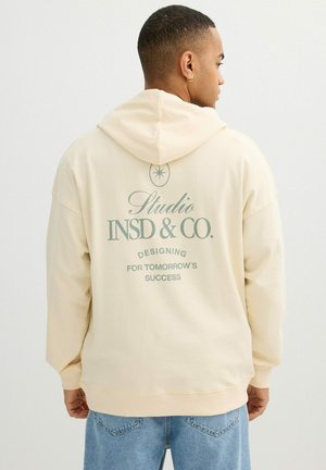 Hombre con sudadera con capucha color crema con el texto "Studio INSD & CO. Designing for Tomorrow's Success" en la espalda, combinado con vaqueros azules.