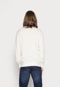 Pullover di colore crema con maniche lunghe, polsini e orlo a coste, e una texture a maglia liscia, indossato con jeans blu scuro.