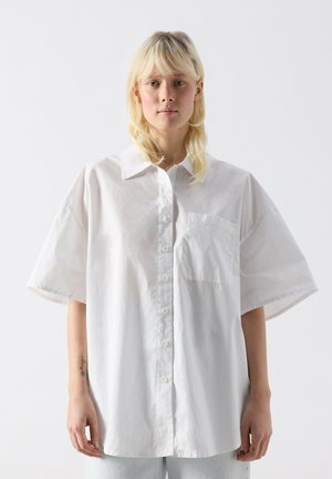 Blonde Person trägt übergroßes weißes Hemd mit kurzen Ärmeln und Brusttasche, steht vor einfachem hellem Hintergrund.