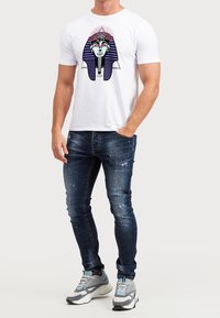 Bílé bavlněné tričko s grafikou obličeje fáraona s tmavými pruhy. Kombinováno s opranými modrými džínami a šedými teniskami.