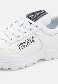 Sapatilhas brancas de atacadores com um cabedal de couro texturizado, apresentando um proeminente logótipo preto "Versace Jeans Couture" e uma sola branca robusta.