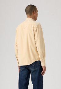 Mann iført en beige langermet skjorte og mørkeblå jeans, som står på siden med ryggen mot kameraet mot en enkel bakgrunn.