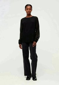 Woman standing wearing black sheer long-sleeve top, black pants with frayed hems, and black shiny shoes on light background.