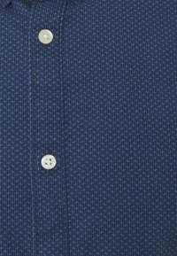 Camicia blu navy con bottoni, caratterizzata da un motivo testurizzato con piccoli puntini blu, rifinita con un bottone rotondo bianco a quattro fori.