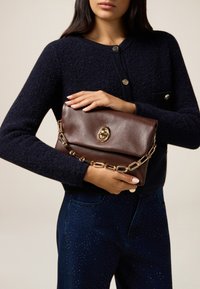 Bolso de piel marrón con una correa de cadena dorada y broche circular, sostenido en las manos, combinado con un suéter texturizado azul marino y jeans oscuros.