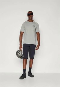 Camiseta de manga corta gris claro, pantalones cortos azul marino, zapatos negros, sosteniendo un casco gris. Los detalles incluyen un logo en la camiseta y en los calcetines.